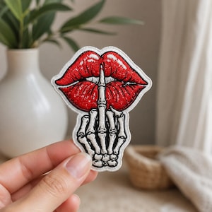 May include: Embroidered patch featuring red lips with a finger over them, and a skeleton hand giving the middle finger. The patch has a white border and is likely for clothing or accessories.