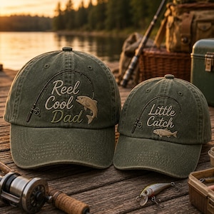 May include: Two olive green baseball caps featuring embroidered fishing rod designs and text. The larger cap displays the words Reel Cool Dad with a fish, while the smaller cap features the words Little Catch with a fish, both in a light tan thread.