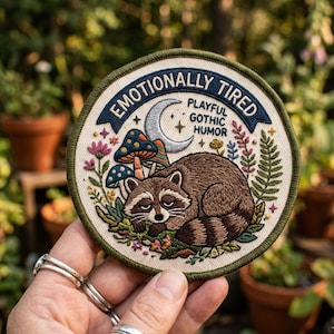 May include: Circular embroidered patch with a raccoon design, mushrooms, and floral elements. The patch includes the text "Emotionally Tired, Playful Gothic Humor". The patch has a green border and is held in a hand.