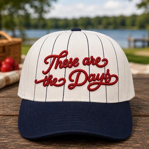 Peut inclure: Casquette de baseball avec une couronne crème à rayures noires et le texte rouge "These are the Days". La visière est bleu marine. Un bouton bleu marine est sur le dessus de la casquette.