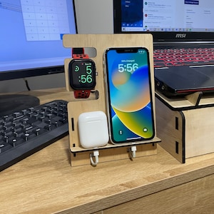 Puede incluir: Un soporte de carga de madera con un Apple Watch negro, una almohadilla de carga inalámbrica blanca y un iPhone negro que muestra 5:56 y 39% de carga. El soporte está sobre un escritorio de madera.