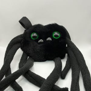 May include: A black plush spider toy with eight long, curved legs. The spider has a fuzzy body and large, round green eyes. The toy is designed to resemble a spider.