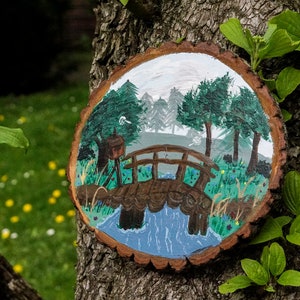 Puede incluir: Una rodaja de madera pintada a mano con una escena caprichosa de un pequeño puente de madera sobre un arroyo azul, rodeado de árboles verdes y un cielo blanco.