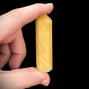 Pu&ograve; includere: Una punta di cristallo giallo dorato tenuta tra le dita su sfondo nero. Il cristallo ha una forma rettangolare con una parte superiore appuntita e motivi interni visibili. Il cristallo &egrave; di un giallo chiaro e traslucido.