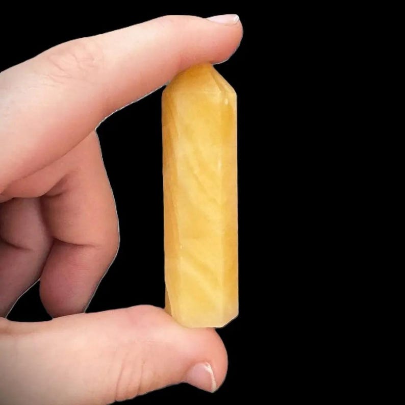 Pu&ograve; includere: Una punta di cristallo giallo dorato tenuta tra le dita su sfondo nero. Il cristallo ha una forma rettangolare con una parte superiore appuntita e motivi interni visibili. Il cristallo &egrave; di un giallo chiaro e traslucido.