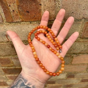Könnte beinhalten: Eine lange Halskette aus orangefarbenen und gelben Perlen. Die Perlen sind rund und glatt und auf einer silbernen Kette aufgereiht.