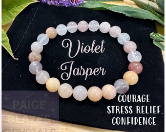 May include: A bracelet made with violet jasper beads. The bracelet is on a black velvet background. The text "Violet Jasper" is written in cursive on the image. The text "Courage, Stress Relief, Confidence" is also written on the image.