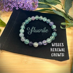 May include: A green and purple fluorite gemstone bracelet on a black velvet pouch. The pouch has the words "Wishes Renewal Growth" printed on it. The bracelet is made of round beads and has the word "Fluorite" written on it.