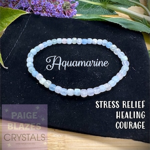 May include: Aquamarine bracelet made with light blue gemstone beads. The bracelet is on a black velvet background. The text "Aquamarine" is written in white on the background. The text "Stress Relief, Healing, Courage" is written in white on the background.