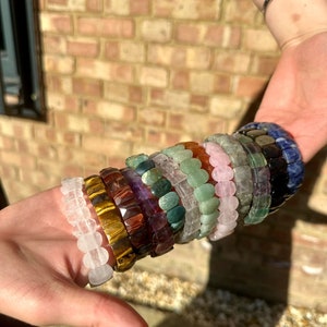 Può includere: Una pila di braccialetti di pietre preziose colorate, tra cui quarzo trasparente, occhio di tigre, ametista, aventurina verde, quarzo rosa e lapislazzuli blu.