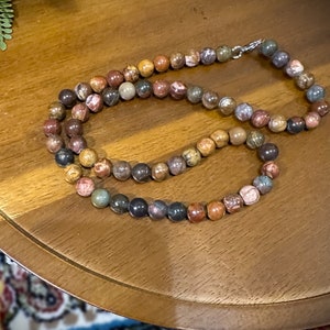 May include: A multi-coloured beaded necklace made of round, polished stones. The necklace is made of various shades of brown, red, and grey.