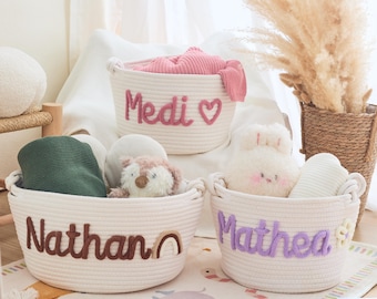 May include: Three off-white rope baskets with embroidered names and contents. The basket labeled "Medi" has pink items, "Nathan" has a green blanket and toys, and "Mathea" has a stuffed bunny. The baskets are on a patterned rug.