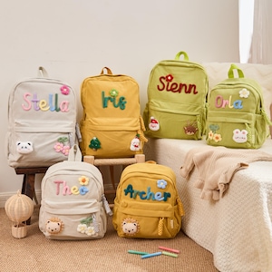 Peut inclure: Plusieurs sacs à dos en velours côtelé de différentes couleurs, chacun personnalisé avec un nom en lettres brodées colorées. Les sacs à dos présentent des appliqués décoratifs tels que des fleurs, des animaux et des ornements festifs.
