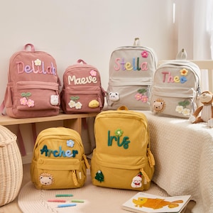 Puede incluir: Una colección de mochilas de pana en varios colores, cada una personalizada con un nombre bordado en colores. Las mochilas tienen aplicaciones florales y de animales decorativas, un bolsillo delantero y un asa superior. Las mochilas son para niños.
