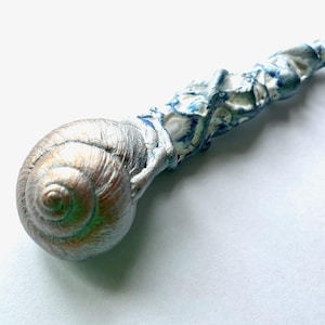 Könnte beinhalten: Ein dekorativer Zauberstab mit Schneckenhaus-Design. Die Schale ist metallisch silberfarben mit grünen Akzenten, und der Griff ist weiß mit blauer Farbe. Ein einzigartiger, handgefertigter Artikel.