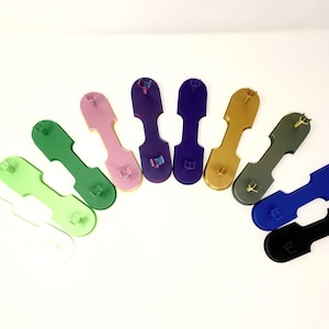 Könnte beinhalten: Eine Sammlung bunter Schlüsselanhänger aus Kunststoff in verschiedenen Farben, darunter Grün, Rosa, Lila, Gold, Grau, Blau und Schwarz. Jeder Schlüsselanhänger hat eine einzigartige Form mit einem kleinen, erhabenen Design.