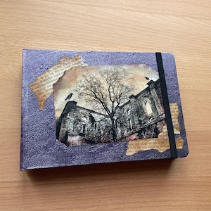 Könnte beinhalten: Ein lila Notizbuch mit strukturierter Hülle, mit einer Collage eines gotischen Gebäudes mit einem Baum und zwei Krähen. Zerrissene Papierränder und ein schwarzes Gummiband vervollständigen das Design.