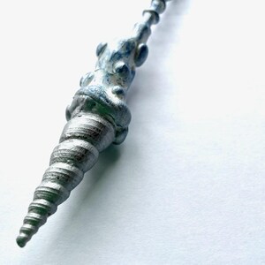 Könnte beinhalten: Ein silberfarbenes, spiralförmiges stabähnliches Objekt mit strukturierter Oberfläche und blauen Akzenten. Der Stab hat ein detailliertes, verziertes Design, das sich zu einer scharfen Spitze verjüngt. Das Objekt befindet sich vor einem weißen Hintergrund.