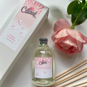 Puede incluir: Una botella de vidrio transparente con una etiqueta rosa y blanca que dice "Cherish Pink Fizz & Grapefruit Reed Diffuser". La botella está sobre una superficie blanca con una rosa rosa y palitos de madera.