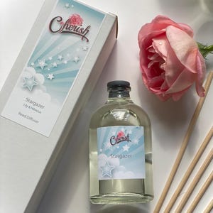 May include: A clear glass bottle with a white label that says "Cherish Stargazer" and a blue and white design with stars. The bottle is filled with a clear liquid and has a black cap. The bottle is sitting on a white surface next to a box with a white label that says "Cherish Stargazer Lily & Hibiscus Reed Diffuser".