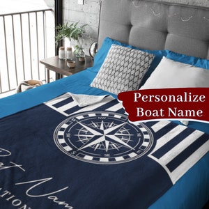 Puede incluir: Una manta de franela a rayas azules y blancas con un diseño de brújula y el texto "Nombre del barco" en blanco. La manta está doblada y se muestra en una cama.