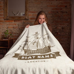 以下が含まれることがあります： 茶色の船のイラストと「BOAT NAME - LOCATION」という文字が印刷された白い毛布です。