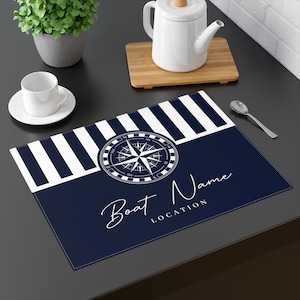 May include: A blue and white striped placemat with a compass design and the text "Boat Name LOCATION".