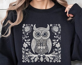 Puede incluir: Sudadera negra con un diseño detallado de búho en escala de grises. El búho está posado en una rama, rodeado de elementos florales y hojas. El diseño está centrado en la parte delantera de la sudadera.