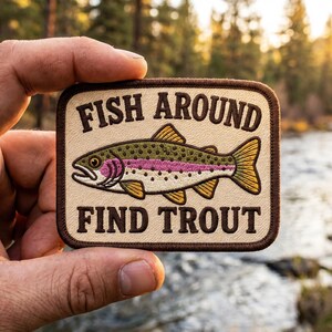 May include: A rectangular embroidered patch with a cream-colored background and a dark brown border. The patch features a detailed illustration of a rainbow trout with green, pink, and yellow scales, accompanied by the text FISH AROUND FIND TROUT.
