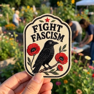 May include: An embroidered patch with an off-white background and a black border. It features a black crow perched on a branch with red poppy flowers. Above the crow, the text FIGHT FASCISM is embroidered in black, with a small red star above it.