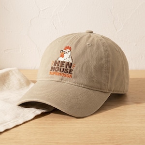 May include: A khaki baseball cap with the text "HEN HOUSE SUPERVISOR" in brown and orange. The cap features an embroidered cartoon chicken wearing glasses. The cap is sitting on a wooden surface next to a white cloth.