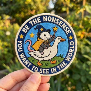 May include: A circular embroidered patch featuring a light blue background with a green grassy base. A gray opossum wearing a yellow cape and blue wizard hat rides a white goose while holding a yellow star wand. The text reads BE THE NONSENSE YOU WANT TO SEE IN THE WORLD.