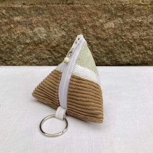 Könnte beinhalten: Ein kleiner, dreieckiger Münzbeutel mit weißem Reißverschluss und silbernem Schlüsselring. Der Beutel besteht aus Cord und gemustertem Stoff in Braun-, Weiß- und Hellgrüntönen. Der Reißverschluss hat eine kleine, weiße Perle.