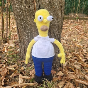 May include: A crocheted yellow character with blue trousers and black shoes. The character has a white shirt and a yellow head with large eyes and a moustache. The character is standing in front of a tree trunk.
