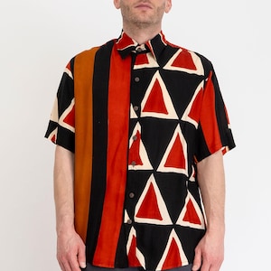 May include: A short-sleeved button-down shirt featuring a split design. The left side displays a black and red geometric triangle pattern on a black background, while the right side features vertical stripes in shades of black, orange, and red.