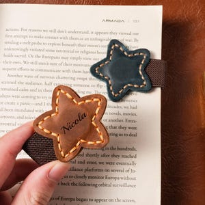 Puede incluir: Dos marcapáginas en forma de estrella con bandas elásticas. Uno es marrón con el nombre "Nicola" y el otro azul oscuro. Ambos tienen bordes cosidos y están en una página de libro.