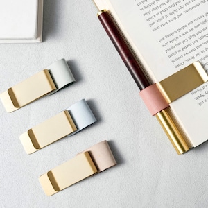 May include: A collection of three metal page clips with colored leather accents in beige, light blue, and pale pink. A pen with a dark wood barrel and gold accents is inserted into a book, held by a similar clip.