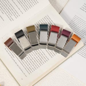 May include: A collection of seven money clips arranged in a fan shape. Each clip features a brushed silver-tone metal body paired with a leather strap in various colors including brown, black, olive, dark blue, red, burgundy, and tan.