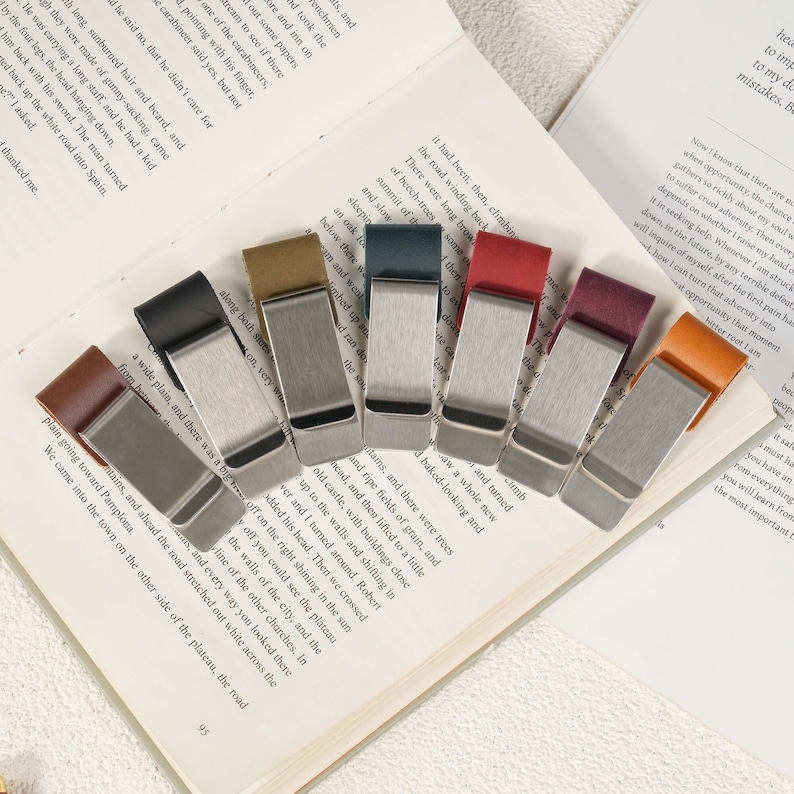 May include: A collection of seven money clips arranged in a fan shape. Each clip features a brushed silver-tone metal body paired with a leather strap in various colors including brown, black, olive, dark blue, red, burgundy, and tan.