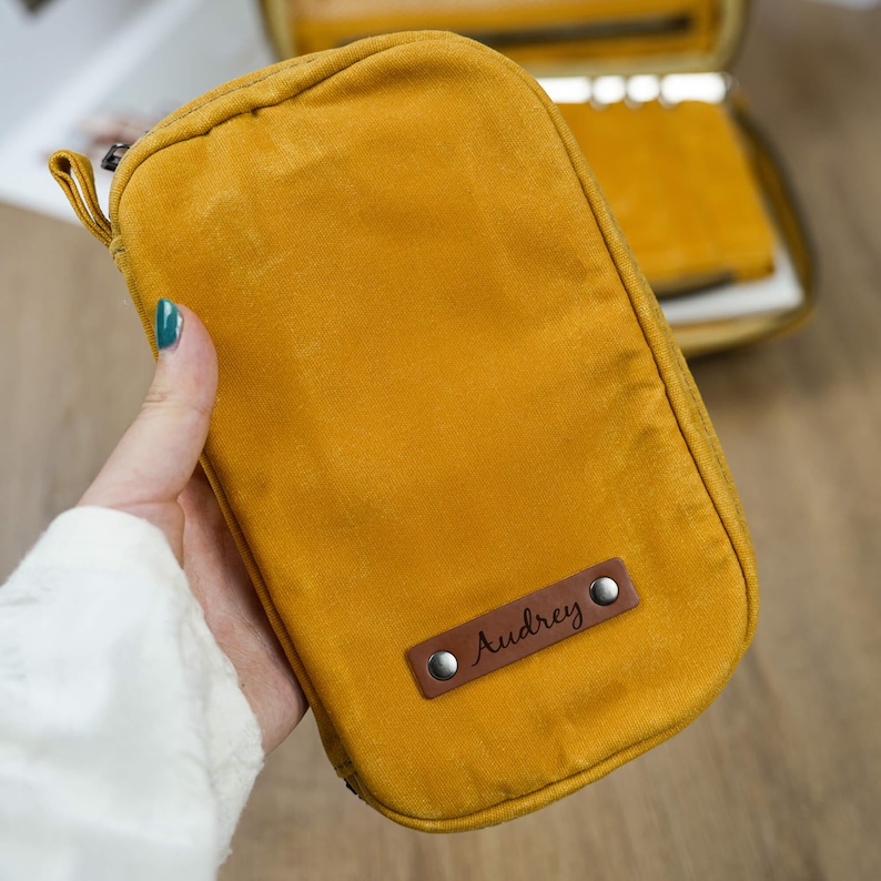 May include: Mustard-yellow rectangular pouch with a zipper closure. A small brown leather tag is attached, with the name "Audrey" engraved. The pouch is held in a hand, with a white sleeve visible in the background.