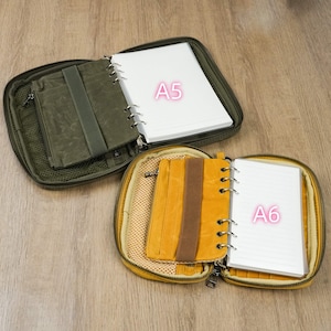 May include: Two open zippered cases, one olive green and one mustard yellow, each containing a notebook. The olive case holds an A5 notebook, and the yellow case holds an A6 notebook. Both cases have interior pockets and elastic bands.