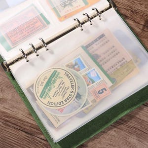 May include: Green binder with clear plastic sleeves filled with vintage ephemera. The binder contains tickets, stamps, and circular stickers with text. The binder is open, revealing the contents, and is resting on a wooden surface.