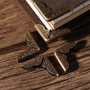 May include: Four ornate metal corner protectors with a decorative, swirling design. The protectors are a dark bronze color and have a vintage aesthetic.