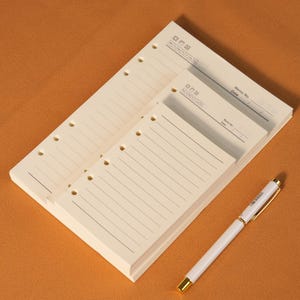 Pode incluir: Um bloco de notas creme com linhas e furos pré-perfurados, juntamente com um bloco menor e uma caneta branca com detalhes dourados. O bloco de notas tem uma seção para "Memo No" e "Data". O fundo é laranja-castanho sólido.