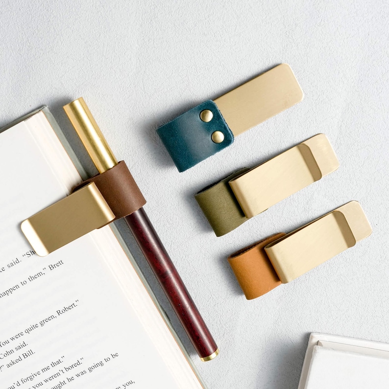 May include: A collection of brass and leather pen holders and page markers. The pen holder features a dark wood pen with a brass tip and a brown leather wrap. Three page markers in green, tan, and teal leather are also shown.