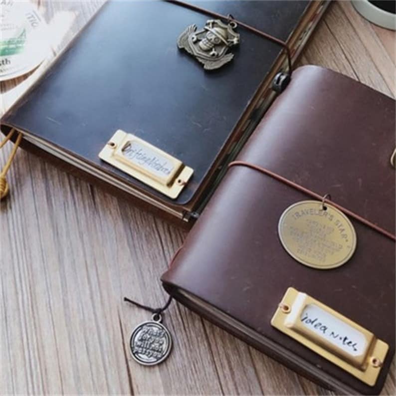 May include: Two brown leather journals with brass accents. One journal has a brass tag that says "Travel's Plan" and the other has a brass tag that says "Idea Notes".