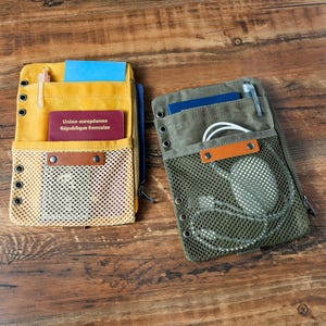 May include: Two canvas travel organisers, one yellow and one olive green, with mesh pockets and metal grommets. The yellow organiser holds a passport with French text. The green organiser contains headphones. Both have zippers and leather accents.