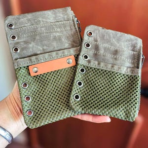 May include: Two olive green canvas and mesh zippered pouches. The top section is a solid olive green canvas with metal grommets. The bottom section is a mesh material. An orange leather tag is attached to the front of one pouch.