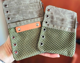Op de afbeelding: Twee olijfgroene canvas en mesh ritszakjes. Het bovenste gedeelte is effen olijfgroen canvas met metalen ogen. Het onderste gedeelte is een mesh materiaal. Een oranje leren label is aan de voorkant van een zakje bevestigd.