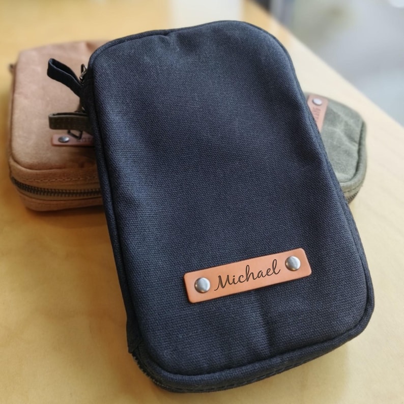 May include: A black canvas zippered pouch with a leather name tag that reads "Michael". The pouch is rectangular and has a rounded top. A second pouch in tan is visible in the background, along with a green pouch.